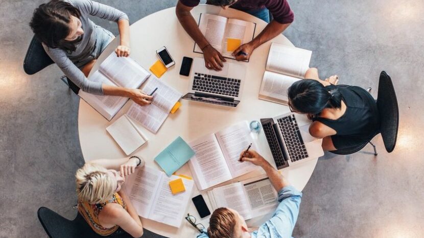 There are 5 students sitting at a small round table with books and laptops. They look like they are doing group study. The view of the picture is from the top looking down at the group of students.