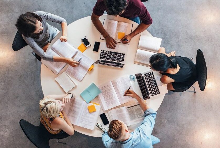 There are 5 students sitting at a small round table with books and laptops. They look like they are doing group study. The view of the picture is from the top looking down at the group of students.